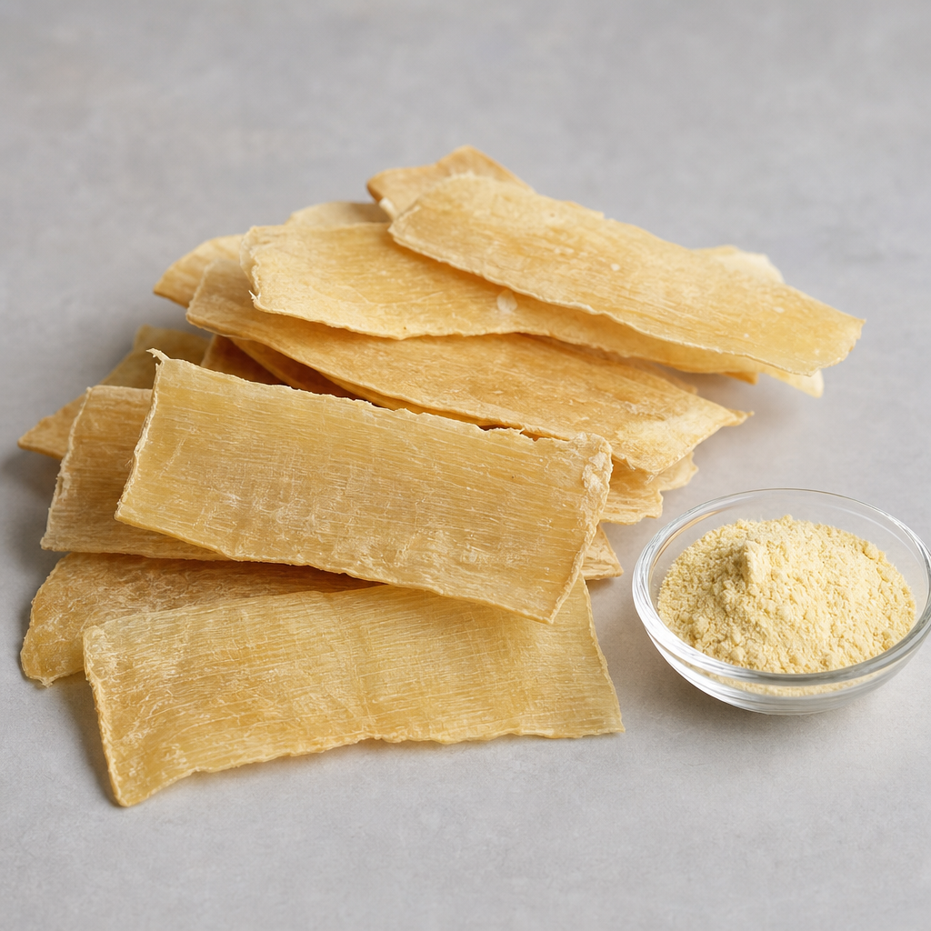 Thin flat dried pale cream to light amber raw-material slices with a matte dry surface stacked in an overlapping pile beside a small glass dish of fine light-yellow powder on a neutral light-grey surface
