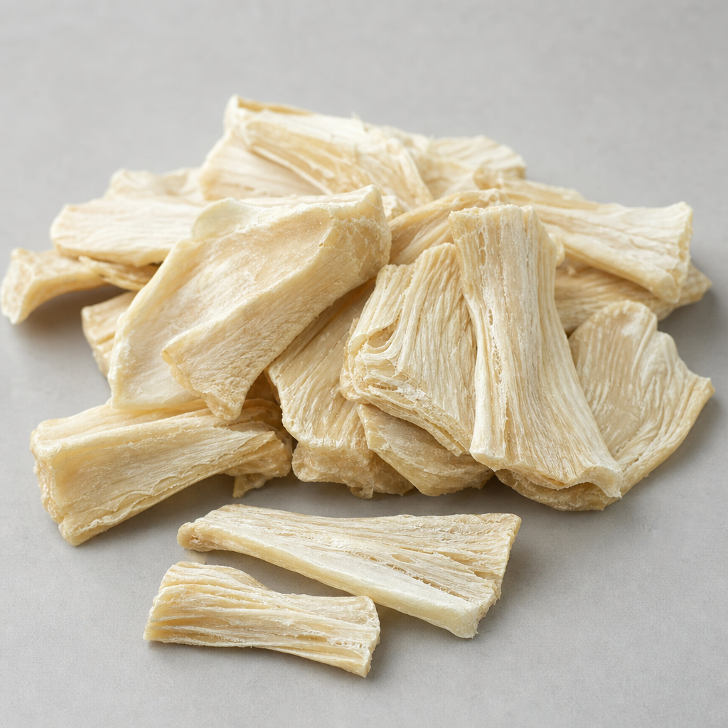Flat irregular pieces of dried pale cream chicken sternal cartilage with a matte dry fibrous surface, arranged in a neat pile with a few pieces showing their layered structure on a neutral light-grey surface